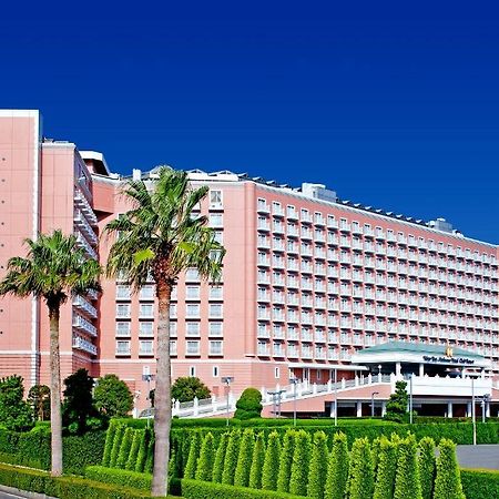 Отель Grand Nikko Tokyo Bay Maihama Ураясу Экстерьер фото The image shows a large, multi-story hotel building featuring a light pink facade. The architecture appears modern, with numerous windows arranged in a grid pattern. In the foreground, there are neatly trimmed shrubs and palm trees, adding a tropical