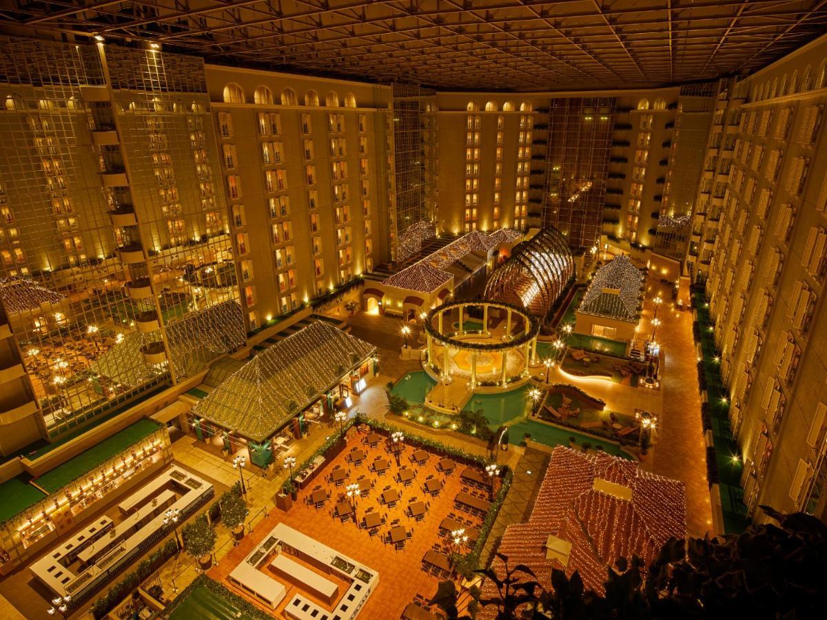 Отель Grand Nikko Tokyo Bay Maihama Ураясу Экстерьер фото The photo shows a large, illuminated atrium of a hotel or resort complex. Several floors of what appear to be guest rooms or apartments surround the central area. The atrium contains a variety of amenities, including a pool or water feature, landscap