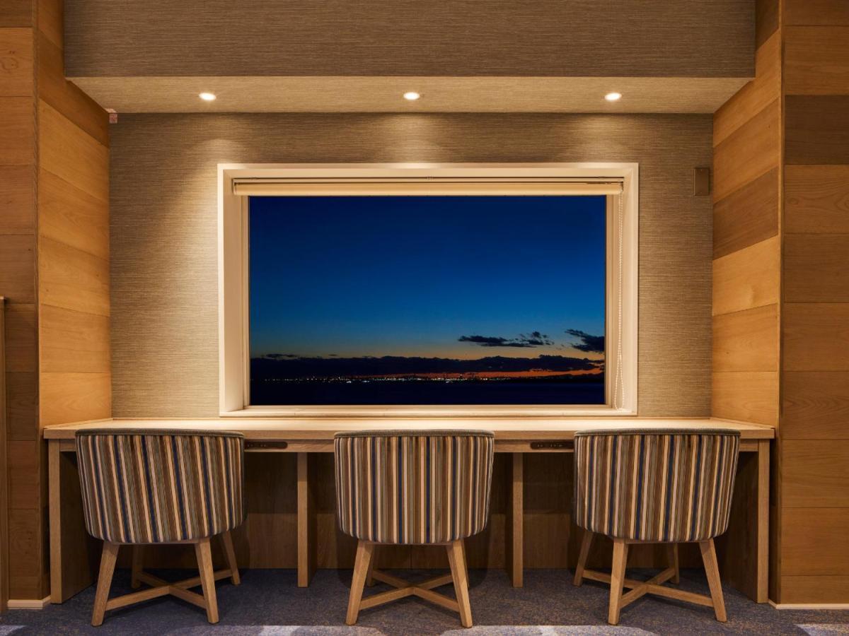 Отель Grand Nikko Tokyo Bay Maihama Ураясу Экстерьер фото The photo depicts a cozy workspace or study area featuring a long, minimalist desk with three chairs. The wall behind the desk is made of wood, creating a warm atmosphere. At the center, there is a large window that showcases a beautiful evening view