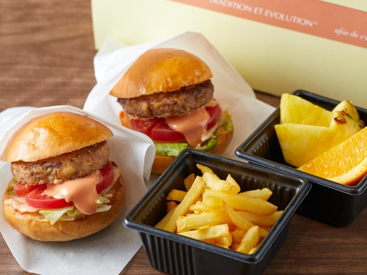 Отель Grand Nikko Tokyo Bay Maihama Ураясу Экстерьер фото The photo shows two small burgers placed on a white background, each with a golden-brown bun, a fried patty, lettuce, tomato, and a creamy sauce. Alongside the burgers, there are two containers: one holding a serving of french fries and the other con