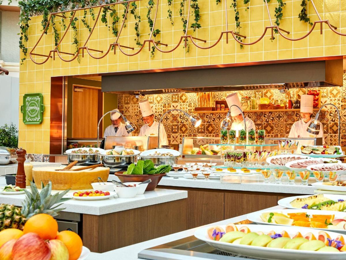 Отель Grand Nikko Tokyo Bay Maihama Ураясу Экстерьер фото The photo shows a vibrant and colorful buffet setup in a restaurant. In the foreground, there are various dishes arranged on white platters, featuring an assortment of fruits, desserts, and other items. The background features chefs in white uniforms