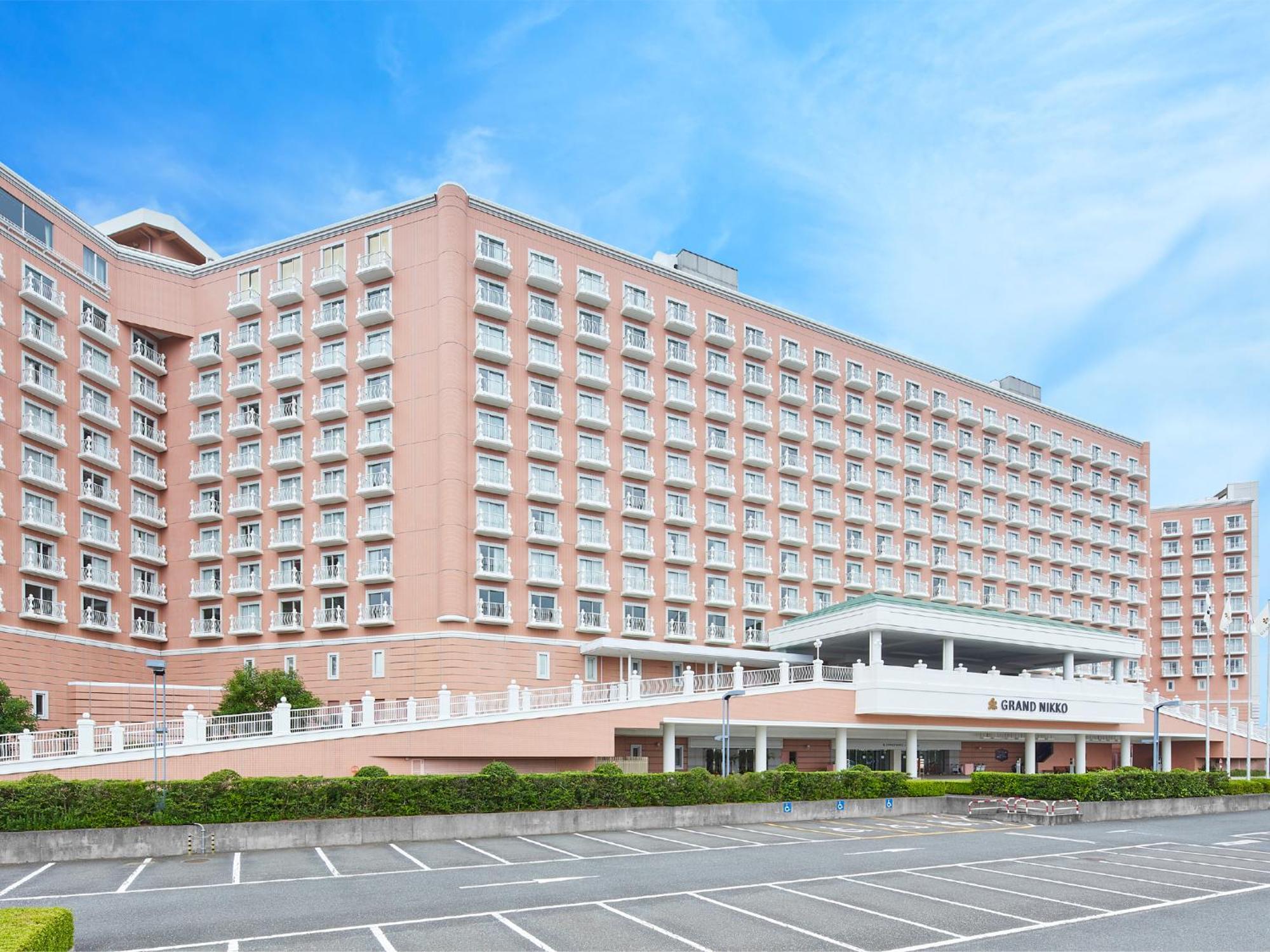 Отель Grand Nikko Tokyo Bay Maihama Ураясу Экстерьер фото The photo shows a large, multi-story hotel building, characterized by its pink facade and modern architectural design. The structure features numerous windows and a spacious entrance area, complemented by a canopy. In front of the hotel, there is a p