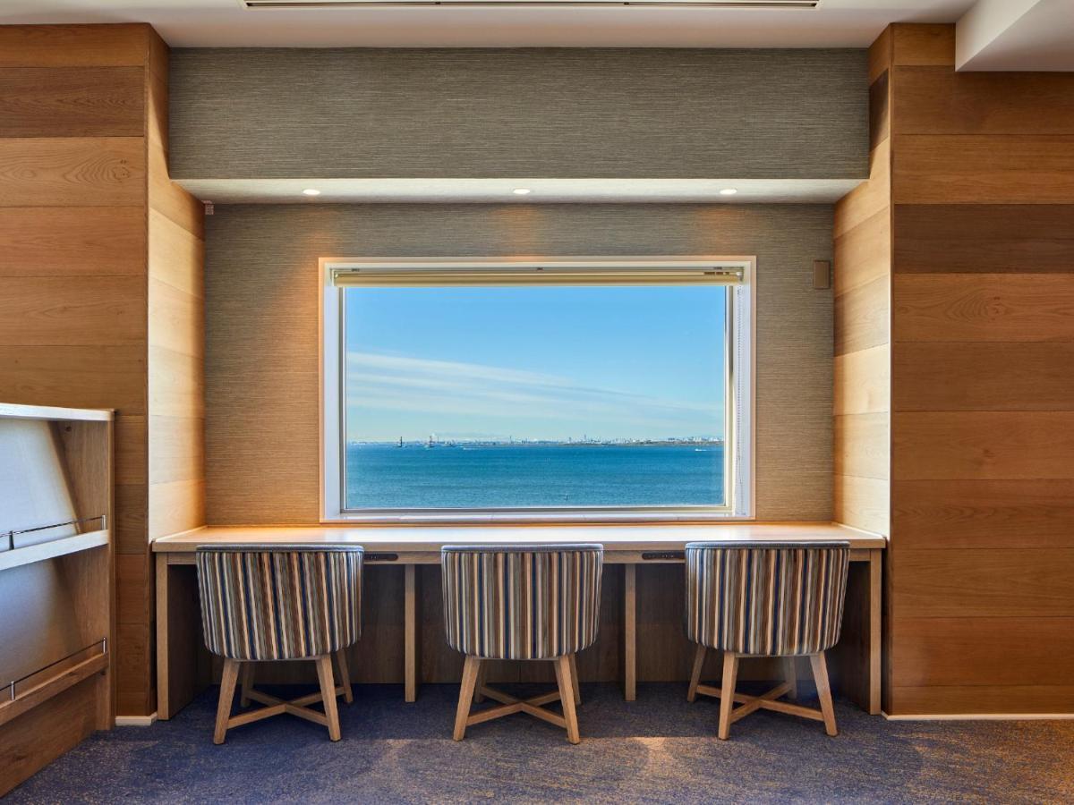 Отель Grand Nikko Tokyo Bay Maihama Ураясу Экстерьер фото The photo shows a modern workspace with three striped chairs positioned at a long, white desk. The desk faces a large window with a view of a tranquil body of water and a distant skyline. The walls around the workspace are made of wood paneling, crea
