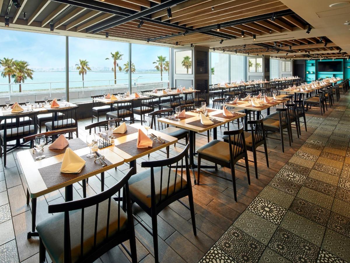 Отель Grand Nikko Tokyo Bay Maihama Ураясу Экстерьер фото The photo shows a modern restaurant interior featuring rows of neatly arranged tables and chairs. The large windows provide a scenic view of palm trees and what appears to be a body of water in the background, suggesting a coastal location. Each tabl