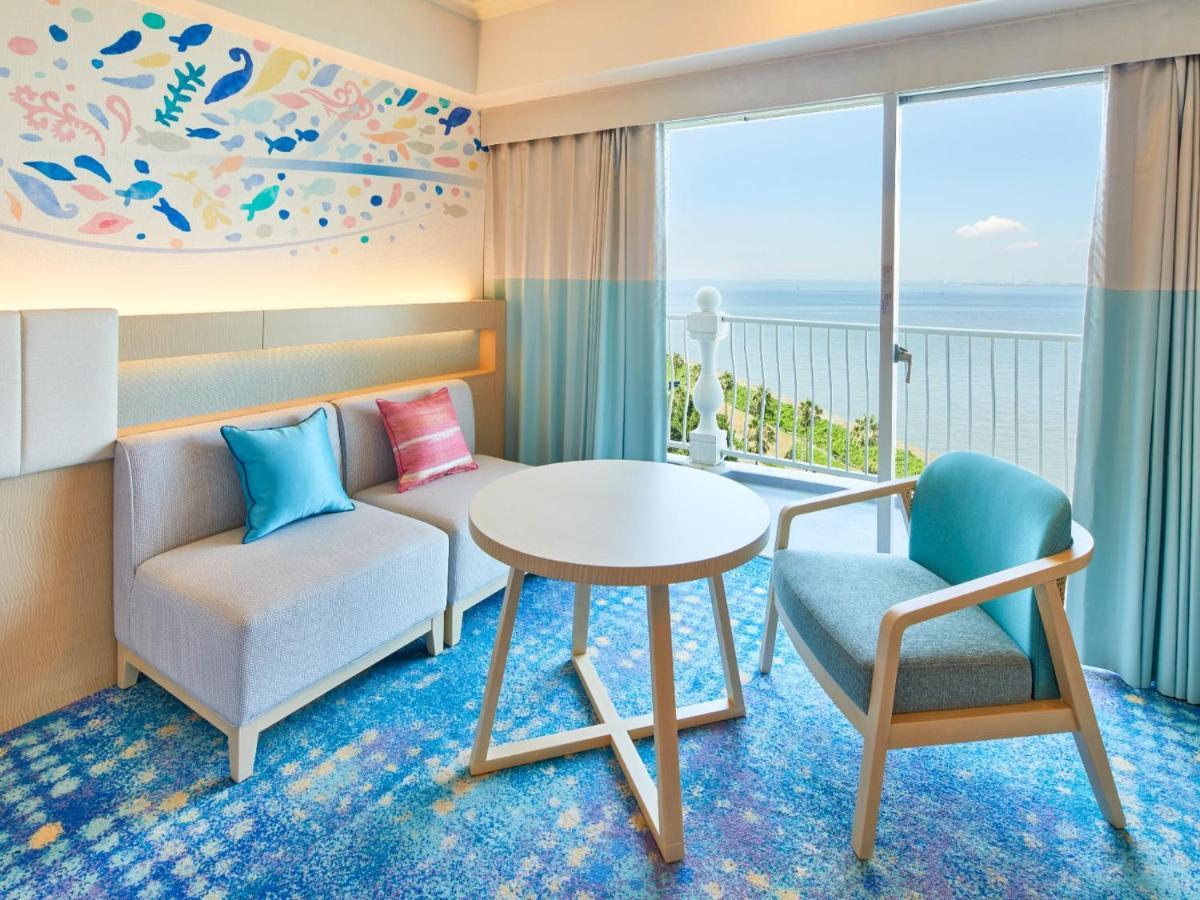 Отель Grand Nikko Tokyo Bay Maihama Ураясу Экстерьер фото The photo shows a modern and stylish interior of a room with a view. There is a cozy seating area featuring a light-colored sofa with decorative cushions in various shades of blue and pink. A round table occupies the center of the space, surrounded b