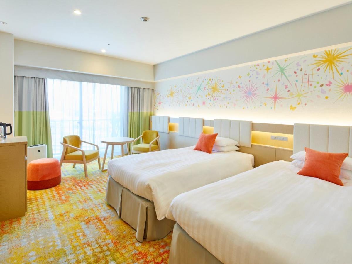 Отель Grand Nikko Tokyo Bay Maihama Ураясу Экстерьер фото The photo shows a modern hotel room featuring two twin beds with light-colored bedding and decorative orange cushions. There is a vibrant, colorful carpet on the floor, and the walls are adorned with playful designs, including bursts of color. A larg