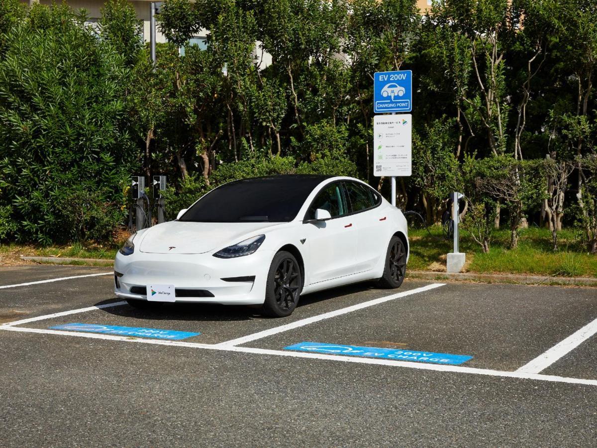 Отель Grand Nikko Tokyo Bay Maihama Ураясу Экстерьер фото The photo shows a white electric car parked in an EV (electric vehicle) charging spot. The car has a sleek design with black wheels and tinted windows. In the background, there are green shrubs and trees, indicating a natural or landscaped environmen