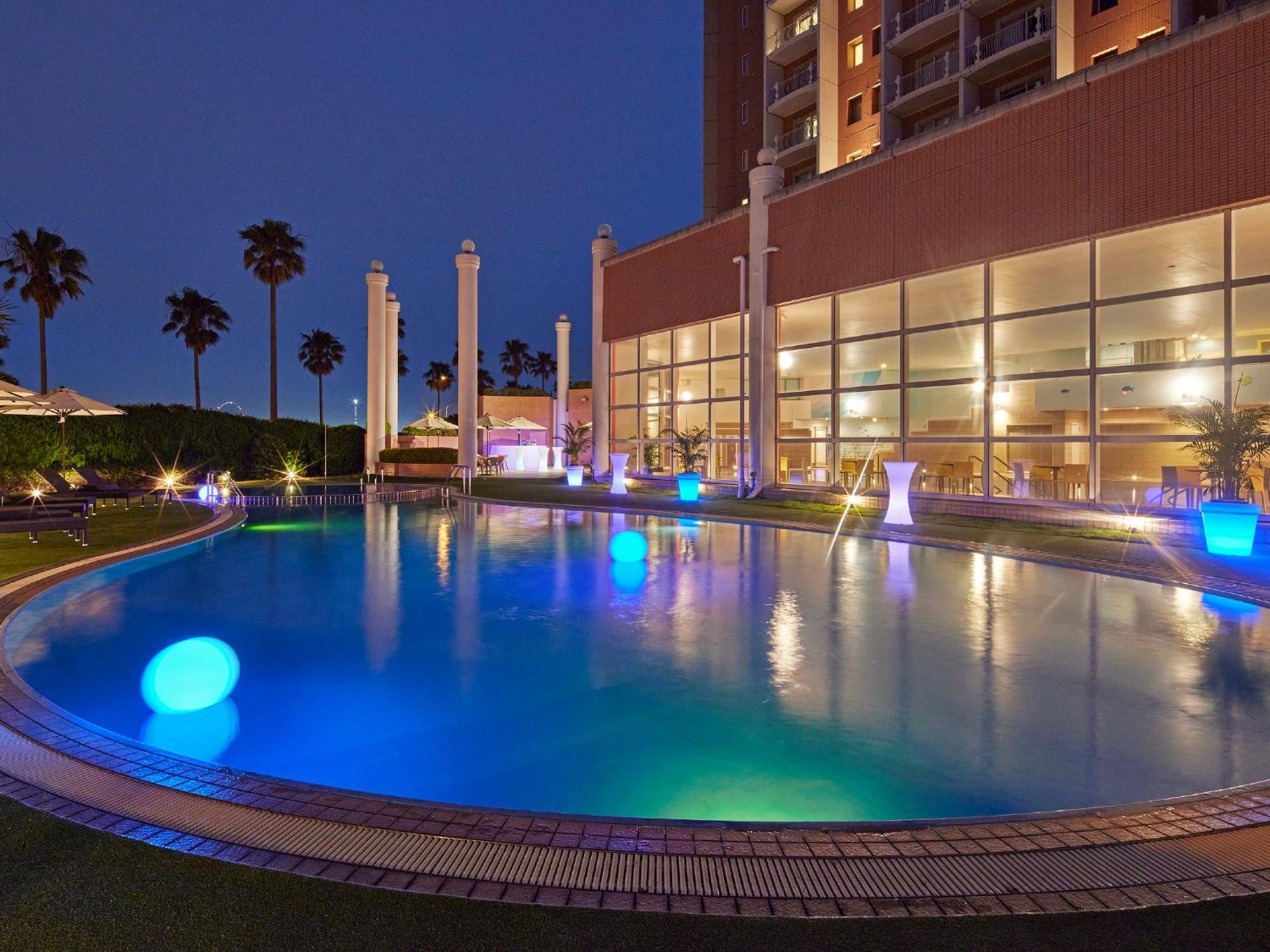 Отель Grand Nikko Tokyo Bay Maihama Ураясу Экстерьер фото The photo shows a well-lit swimming pool at night, surrounded by a lush green area. The pool features glowing blue lights that create a serene and inviting atmosphere. Palm trees can be seen in the background, adding a tropical feel to the scene. Add