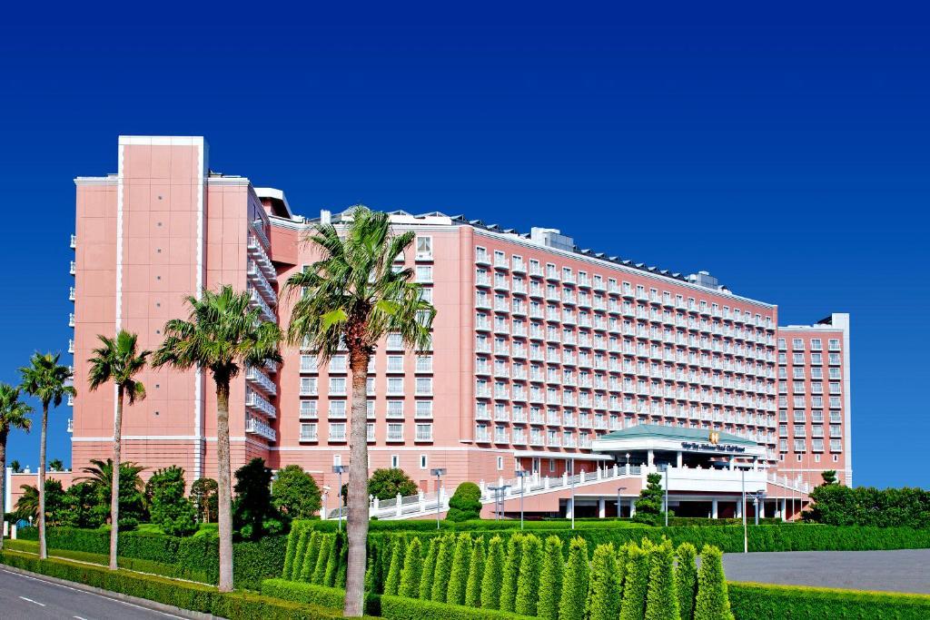 Отель Grand Nikko Tokyo Bay Maihama Ураясу Экстерьер фото The image shows a large, multi-story hotel building featuring a light pink facade. The architecture appears modern, with numerous windows arranged in a grid pattern. In the foreground, there are neatly trimmed shrubs and palm trees, adding a tropical